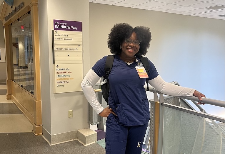 A smiling nursing student in navy scrubs and a backpack stands in a hospital corridor next to a wayfinding sign reading 'You are on Rainbow Way.